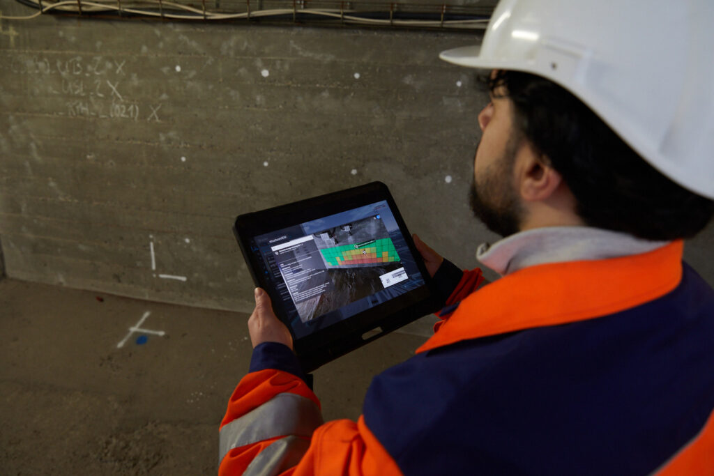 Person mit weißem Schutzhelm und orange-blauer Arbeitskleidung hält ein Tablet mit einer farbigen 3D-Darstellung in einer Baustelle