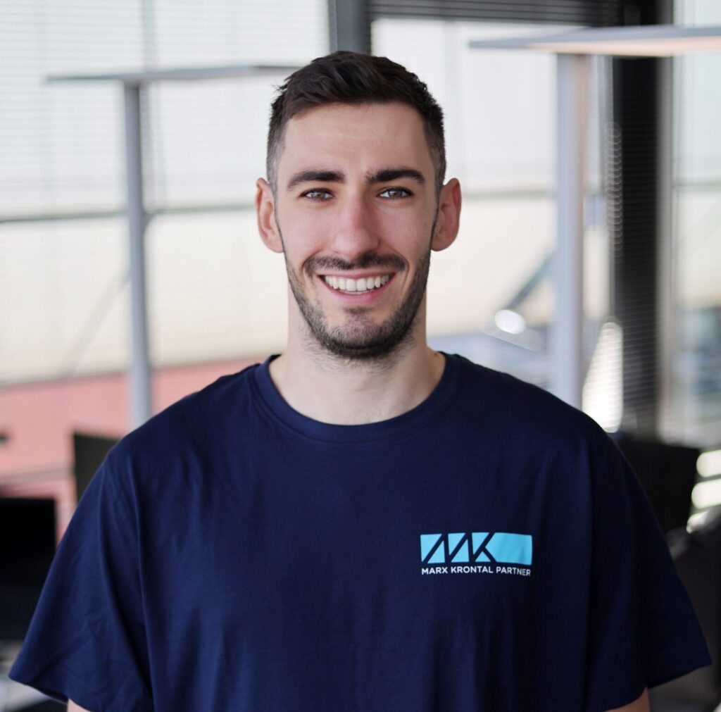 Mann mit kurzem Haar und Bart trägt ein dunkelblaues T-Shirt mit dem Logo und Schriftzug 'MARX KRONTAL PARTNER' vor einem Bürofenster.
