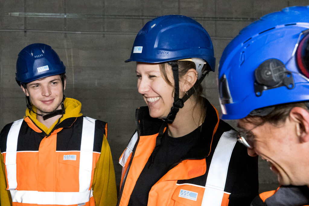 Drei Personen mit blauen Schutzhelmen und orangefarbenen Warnwesten vor einer grauen Wand