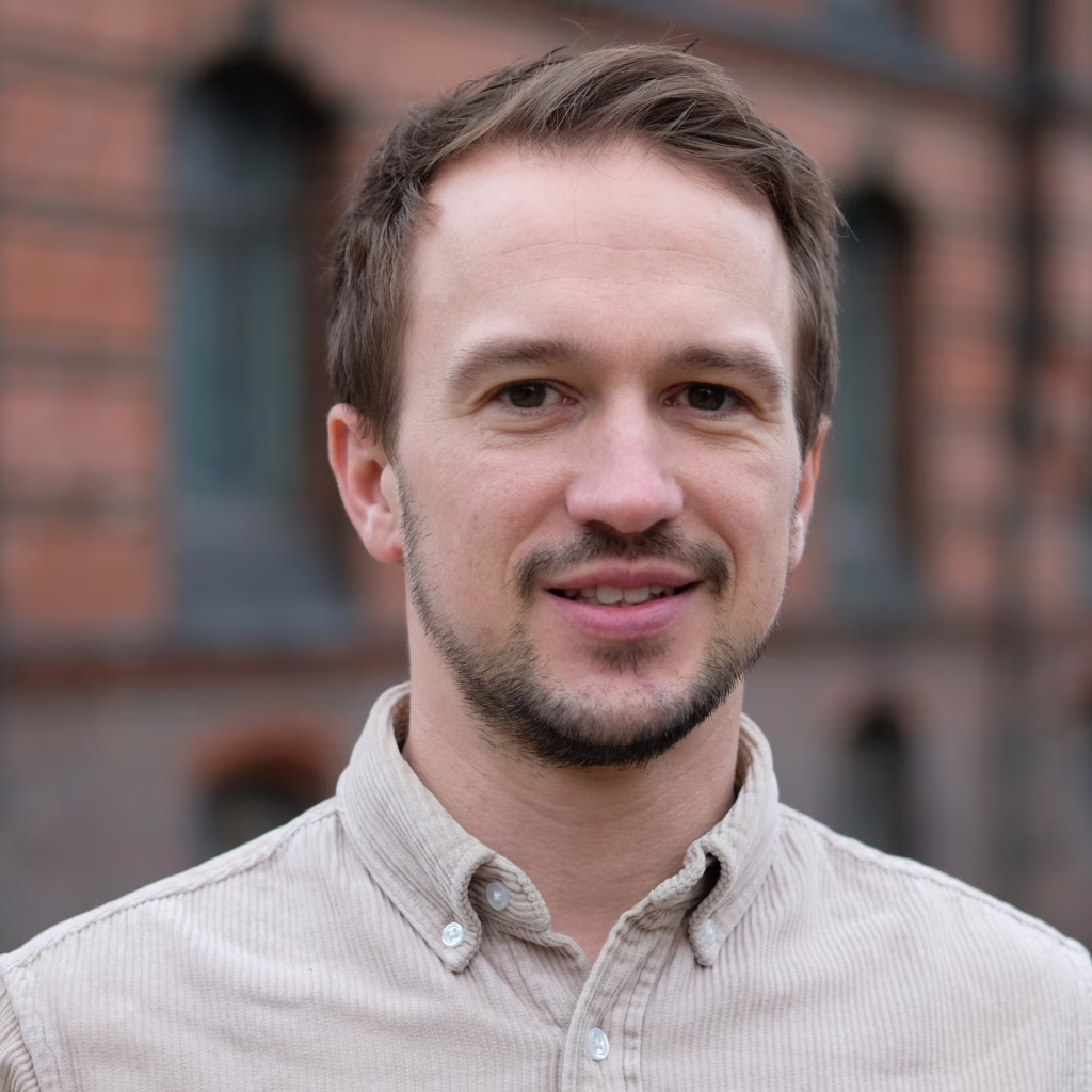 Mann mit kurzem Haar und Hemd mit Knopfleiste vor einer Backsteinwand.