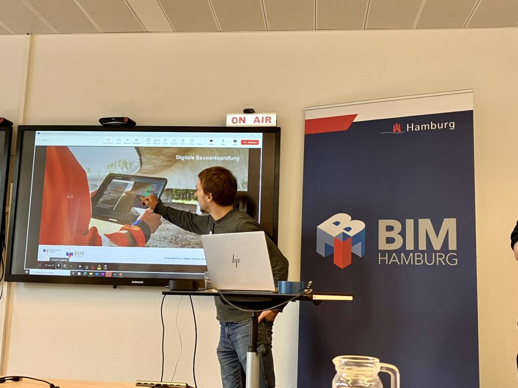 Eine Person zeigt auf einem Bildschirm eine Präsentation zur digitalen Bauwerksprüfung, daneben steht ein Banner mit der Aufschrift 'BIM HAMBURG'.