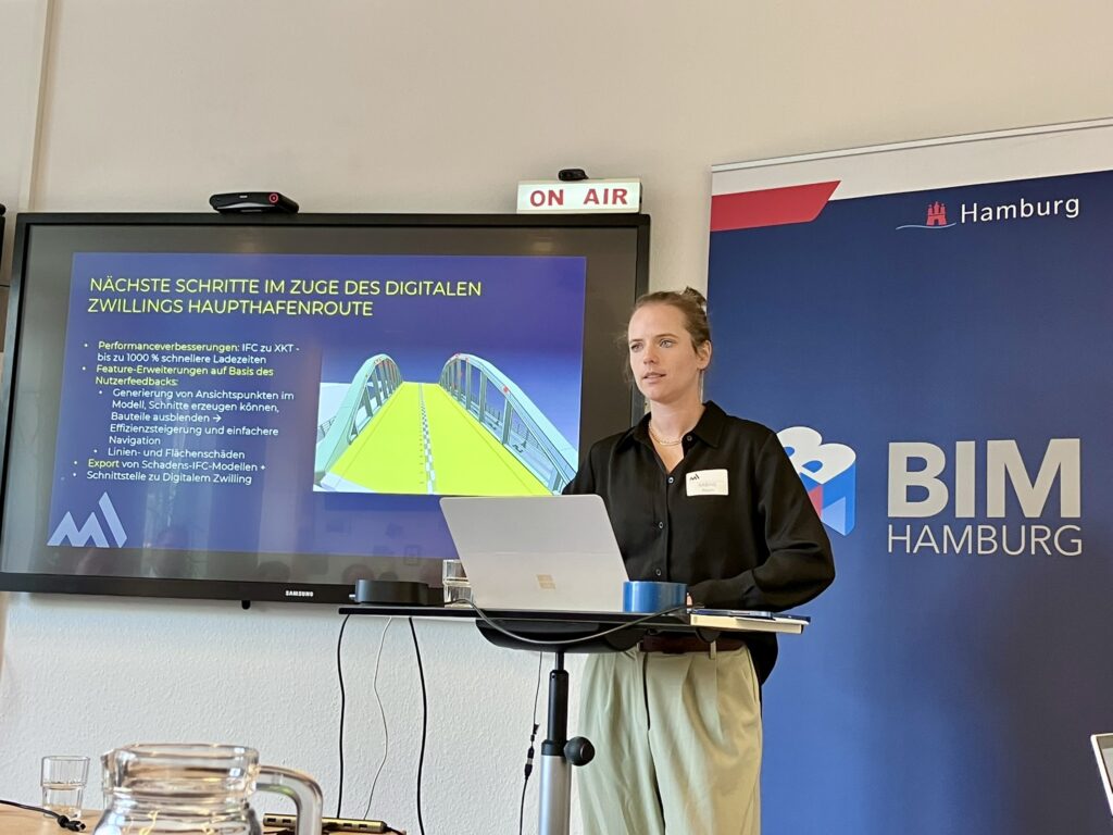Eine Person steht an einem Stehpult mit Laptop vor einem Bildschirm, auf dem eine Präsentation mit dem Titel 'NÄCHSTE SCHRITTE IM ZUGE DES DIGITALEN ZWILLINGS HAUPTFAHRENROUTE' zu sehen ist, daneben ein Banner mit dem Schriftzug 'BIM HAMBURG'.