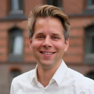 Mann mit hellblondem Haar trägt ein weißes Hemd vor einem unscharfen Backsteingebäude.