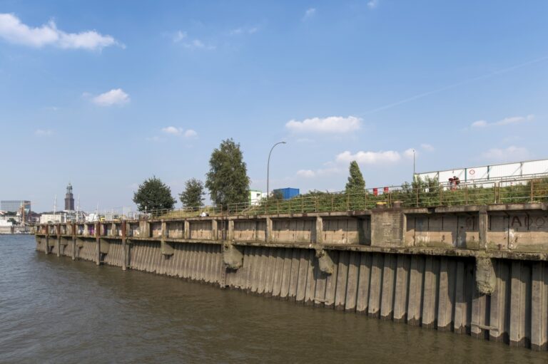 Kaimauer mit Betonpfeilern am Flussufer unter blauem Himmel mit vereinzelten Wolken