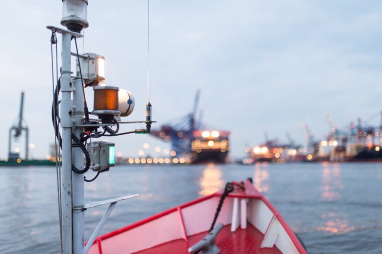 Bug eines Bootes mit rotem Rand und Metallmast im Vordergrund vor unscharfer Hafenkulisse mit beleuchteten Kränen und Schiffen