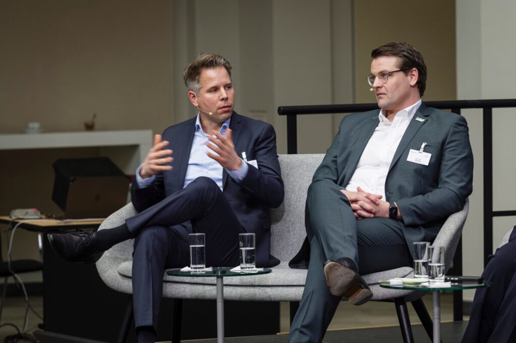 Zwei Männer in Anzügen sitzen auf einem Sofa und führen eine Diskussion, vor ihnen stehen Gläser mit Wasser auf kleinen Tischen