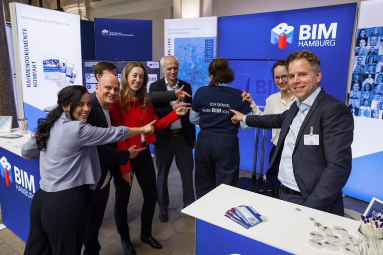 Gruppe von Personen steht vor einem Messestand mit mehreren Bildschirmen und Bannern, auf einem Rücken ist der Schriftzug 'ICH DENKE, ALSO BIM ICH' zu sehen