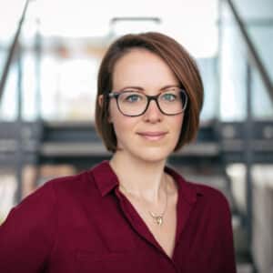 Person mit schulterlangem braunem Haar und rotem Hemd vor unscharfem Treppenhaus