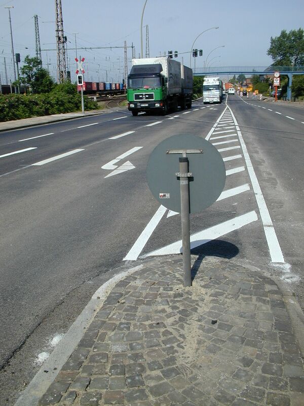 Verkehrsinsel mit rundem grauem Verkehrsschild ohne sichtbare Beschriftung auf gepflastertem Untergrund an einer mehrspurigen Straße mit Lkw und Autos im Hintergrund