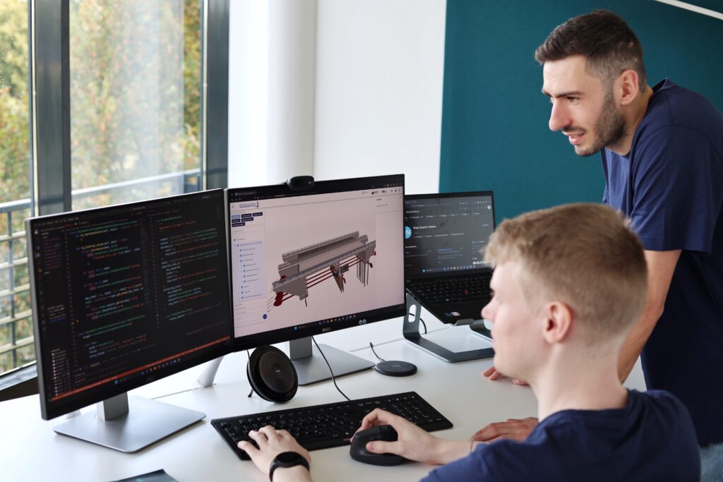 Zwei Personen an einem Schreibtisch mit drei Monitoren, auf denen Programmiercode und ein 3D-Modell eines Bauwerks zu sehen sind