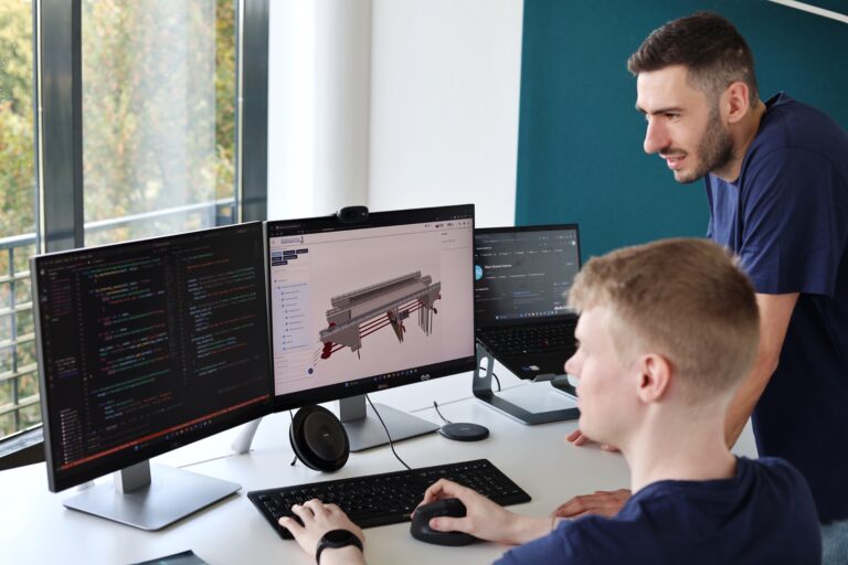 Zwei Personen an einem Schreibtisch mit drei Monitoren, auf denen Programmiercode und ein 3D-Modell eines Bauwerks zu sehen sind