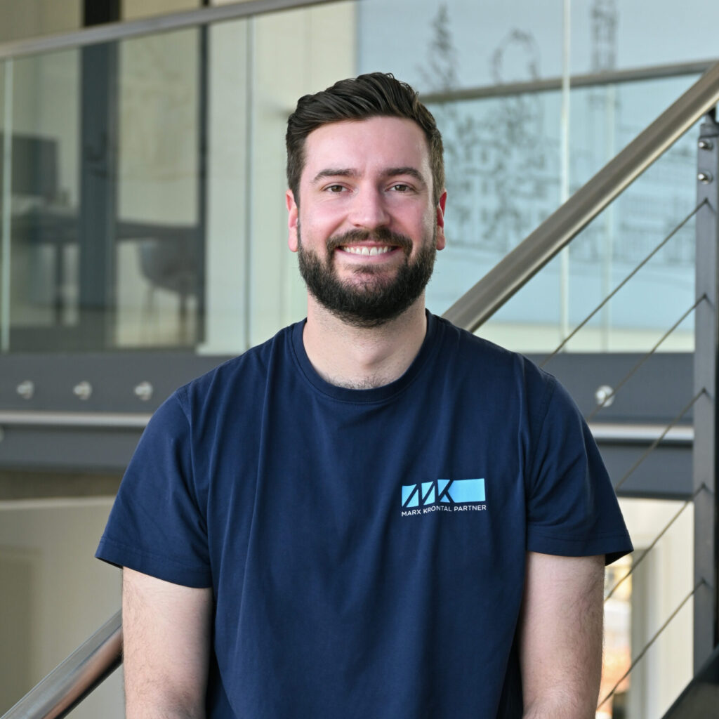 Mann mit dunklem kurz geschnittenem Haar und Bart trägt ein dunkelblaues T-Shirt mit hellblauem Logo und Schriftzug 'MARK KRONTAL PARTNER' vor einer Treppe mit Glasgeländer