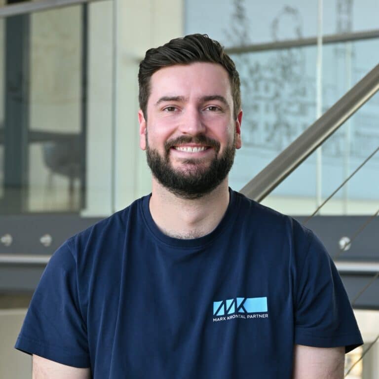 Mann mit dunklem kurz geschnittenem Haar und Bart trägt ein dunkelblaues T-Shirt mit hellblauem Logo und Schriftzug 'MARK KRONTAL PARTNER' vor einer Treppe mit Glasgeländer